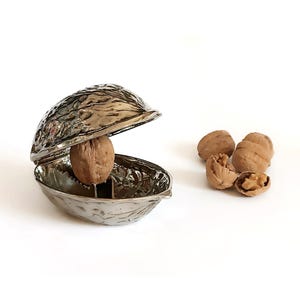 Op de afbeelding: Een zilverkleurige notenkraker in de vorm van een walnootschil, met een walnoot erin. Verschillende hele en gebroken walnoten liggen in de buurt verspreid, alles op een witte achtergrond.