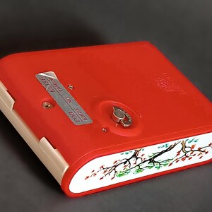 K&ouml;nnte beinhalten: Eine rote Vintage-Zigarettenetui mit einem wei&szlig;en und floralen Design am Rand. Das Etui hat einen silbernen Metallverschluss und der Text "FOR ERIZE NO TAMPER" ist sichtbar.