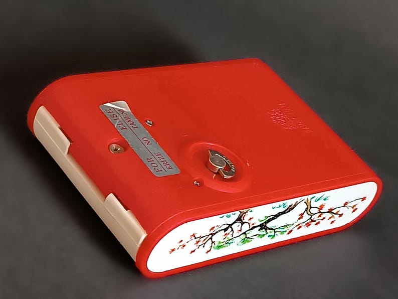 K&ouml;nnte beinhalten: Eine rote Vintage-Zigarettenetui mit einem wei&szlig;en und floralen Design am Rand. Das Etui hat einen silbernen Metallverschluss und der Text "FOR ERIZE NO TAMPER" ist sichtbar.
