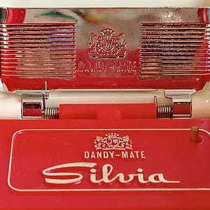 K&ouml;nnte beinhalten: Ein rotes und wei&szlig;es Dandy-Mate Silvia Kompakt-Make-up. Das Kompakt-Make-up hat eine rote Basis mit dem Namen "Silvia" in wei&szlig;er Schrift. Die Oberseite ist verchromt mit den Worten "Dandy-Mate" und einem Wappen. Das Kompakt-Make-up ist klappbar.