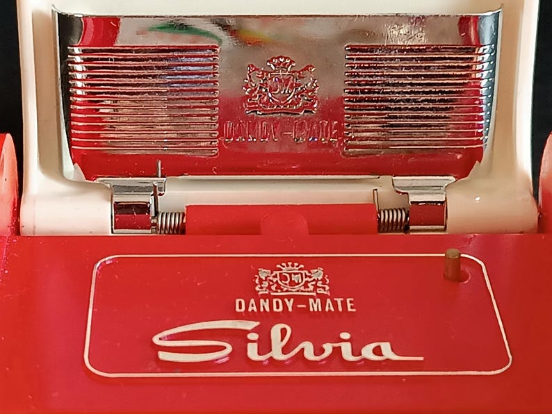 K&ouml;nnte beinhalten: Ein rotes und wei&szlig;es Dandy-Mate Silvia Kompakt-Make-up. Das Kompakt-Make-up hat eine rote Basis mit dem Namen "Silvia" in wei&szlig;er Schrift. Die Oberseite ist verchromt mit den Worten "Dandy-Mate" und einem Wappen. Das Kompakt-Make-up ist klappbar.