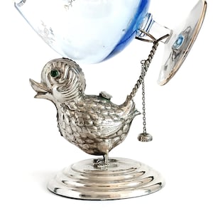 May include: A silver-colored duck-shaped wine glass holder with a blue and clear glass. The duck has green eyes and a chain that connects to the glass. The holder sits on a round, silver-colored base.