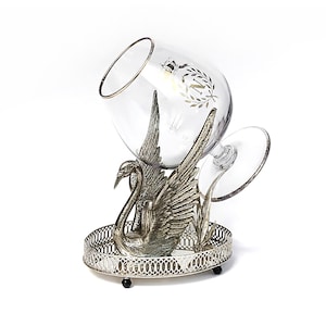 May include: A clear glass brandy snifter with a gold rim, resting in a silver swan-shaped holder. The holder sits on a silver filigree tray. The glass has a gold emblem. The swan has detailed feathers and a long neck.