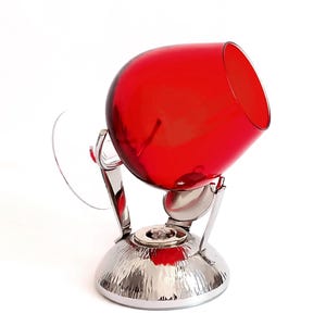 Op de afbeelding: Een rode glazen karaf met een zilveren metalen standaard. De karaf is schuin geplaatst en de standaard heeft een getextureerd oppervlak.