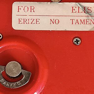K&ouml;nnte beinhalten: Nahaufnahme eines roten Objekts mit einem silbernen Metallzifferblatt und dem Wort "SANKYO". Auf dem Objekt befindet sich ein wei&szlig;es Etikett mit den Worten "FOR ELISE ERIZE NO TAMENI". Zwei Schrauben sind sichtbar.
