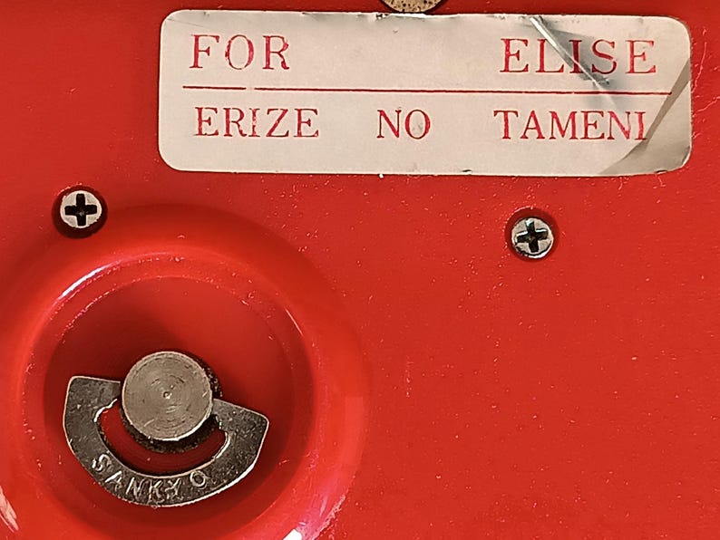 K&ouml;nnte beinhalten: Nahaufnahme eines roten Objekts mit einem silbernen Metallzifferblatt und dem Wort "SANKYO". Auf dem Objekt befindet sich ein wei&szlig;es Etikett mit den Worten "FOR ELISE ERIZE NO TAMENI". Zwei Schrauben sind sichtbar.