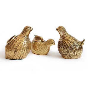 Pode incluir: Um conjunto de três figurinhas de pássaros de metal dourado. As figurinhas têm a forma de codornizes e são usadas como isqueiro, cinzeiro e porta-anéis.
