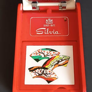K&ouml;nnte beinhalten: Ein rot-wei&szlig;es Dandy-Mate Silvia Zigarettenetui. Das Etui hat eine dekorative Platte mit drei stilisierten F&auml;cherdesigns in Gr&uuml;n, Orange und Gelb. Das Etui ist ge&ouml;ffnet und gibt den Blick ins Innere frei.