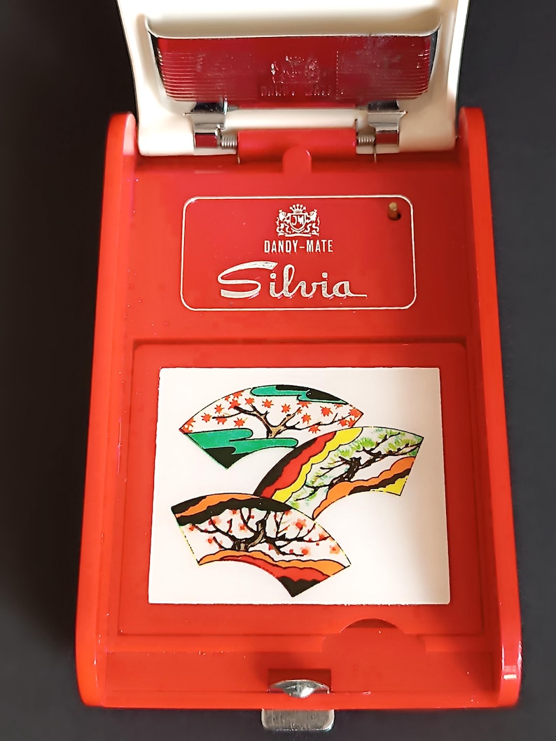 K&ouml;nnte beinhalten: Ein rot-wei&szlig;es Dandy-Mate Silvia Zigarettenetui. Das Etui hat eine dekorative Platte mit drei stilisierten F&auml;cherdesigns in Gr&uuml;n, Orange und Gelb. Das Etui ist ge&ouml;ffnet und gibt den Blick ins Innere frei.