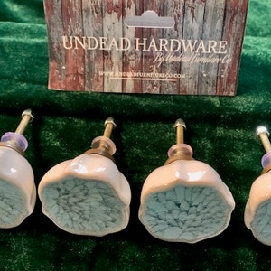 May include: Set of four ceramic cabinet knobs with a light pink base and a blue and white marbled design. The knobs have a gold metal stem and a purple plastic base.