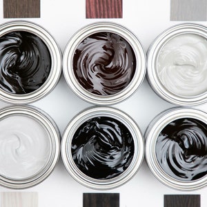 May include: Six open cans of paint with different colours of paint inside. The colours are black, dark brown, light brown, grey, and white. The cans are arranged in two rows of three.