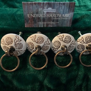 May include: Four white ceramic drawer pulls with a floral design and a brass ring. The pulls are on a green velvet surface. The text "UNDEAD HARDWARE" is visible on a card in the background.
