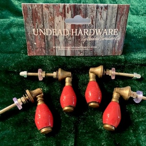 Op de afbeelding: Set van vijf kastknoppen met rode, druppelvormige handgrepen en messingkleurige hardware. De knoppen zijn gerangschikt op een groene fluwelen ondergrond, met een kartonnen bord met de tekst "Undead Hardware".