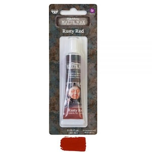 Op de afbeelding: Een tube "Rusty Red" matte wax, 20 ml, wordt getoond op een productkaart. De productkaart heeft een roestige, getextureerde achtergrond. De tube is donker met witte tekst en een gezichtsillustratie. Een staal van de rode wax staat onderaan.
