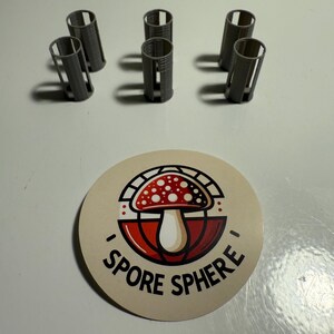 May include: A collection of six gray, cylindrical objects with vertical slits, and a circular sticker. The sticker features a red and white mushroom design with the text "I SPORE SPHERE" in a circular layout.