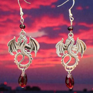 May include: A pair of silver-toned dragon earrings. Each earring features a dragon pendant with outstretched wings, a small red bead, and a teardrop-shaped red gemstone. The earrings hang from a silver hook.