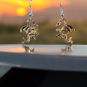 May include: A pair of gold-colored dragon earrings. Each earring features a detailed dragon pendant with outstretched wings, suspended from a silver-colored fish hook. The background shows a sunset with orange and yellow hues.