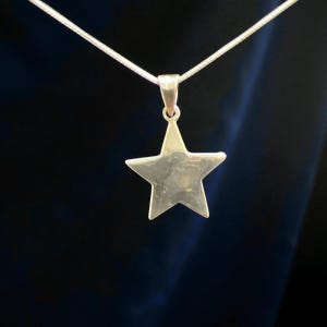 Op de afbeelding: Een zilverkleurige sterhanger ketting. De hanger is een vijfhoekige ster die aan een fijne zilveren ketting hangt. De ketting wordt getoond tegen een donkere achtergrond.