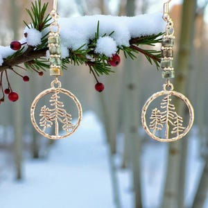 Peut inclure: Boucles d'oreilles dorées avec un motif d'arbre, suspendues à une branche enneigée avec des baies rouges. Les boucles d'oreilles ont un pendentif circulaire avec une silhouette d'arbre découpée et un accent de perles. L'arrière-plan est une scène hivernale floue.