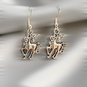 May include: A pair of silver-toned unicorn earrings. Each earring features a detailed unicorn pendant with a floral mane and tail, suspended from a simple hook. The earrings are displayed against a blurred, silvery background.
