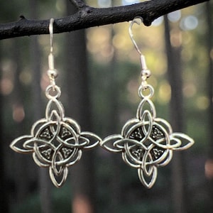 May include: A pair of silver-colored Celtic knot earrings. Each earring features an intricate, symmetrical design with a central knot and four outward-facing lobes. The earrings hang from simple fish hook ear wires, with a blurred forest background.