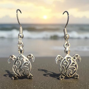 Puede incluir: Un par de pendientes plateados con forma de tortuga. Cada pendiente presenta un diseño detallado de tortuga calada con un patrón decorativo. Los pendientes cuelgan de un gancho simple. El fondo muestra una escena de playa con el océano y una puesta de sol.