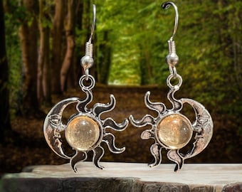 May include: A pair of silver-toned earrings featuring a sun and moon design. Each earring has a central, round, amber-colored stone. The sun has stylized rays, and the crescent moon is detailed with a face. The earrings hang from a simple hook.