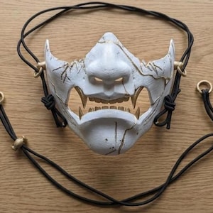May include: A white and gold Oni mask with a fearsome expression, featuring sharp teeth and a cracked texture. The mask has black cords attached for securing it. The mask is a decorative item.