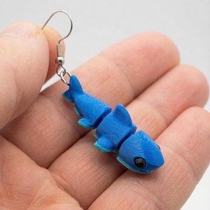 May include: A blue articulated shark earring with a silver hook. The shark is segmented, allowing for movement, and features a light blue accent on the fins and tail. The shark has a black eye.