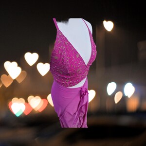 May include: A pink two-piece outfit with a crop top and a wrap skirt. The top has a deep V-neckline and is decorated with gold embroidery. The skirt is made of a soft, flowing fabric and has a tie at the waist.