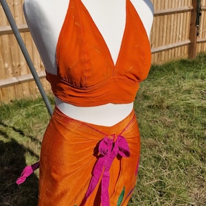 May include: A halter top and wrap skirt set in a bright orange color. The top has a deep V-neckline and the skirt has a pink ribbon tie and floral embroidery.