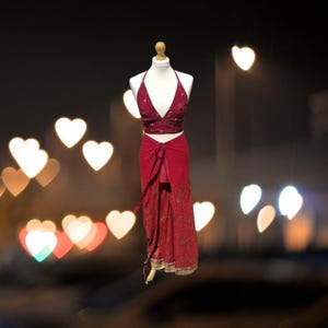 May include: A red halter top and matching wrap skirt with gold embroidery. The outfit is on a mannequin against a blurry background of heart-shaped lights.