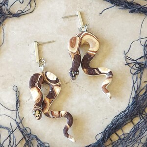 May include: A pair of snake earrings made of polymer clay. The snakes are brown, white, and black with a marbled pattern. The earrings are attached to silver hooks.