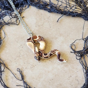 May include: A handcrafted snake pendant necklace. The pendant is made of tan, brown, and cream-colored clay, with a detailed snake design. The necklace has a silver-colored chain. The snake pendant is approximately 3 inches long.
