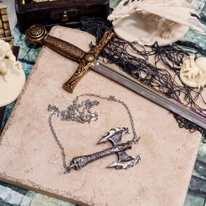 May include: A decorative fantasy-themed image featuring a silver axe pendant necklace, a sword with a gold handle, and a small white dragon figurine. The items are arranged on a textured, light-colored surface, suggesting a collection of fantasy accessories.