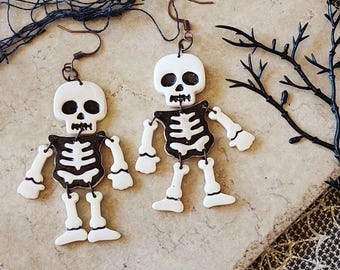 May include: A pair of Halloween-themed earrings featuring white skeleton figures with black rib cages and limbs. The earrings have copper-colored hooks and are set against a neutral-toned background, evoking a spooky yet playful aesthetic.
