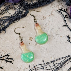 May include: A pair of green and white potion bottle earrings with cork stoppers and gold hooks. The earrings are on a light brown textured surface.