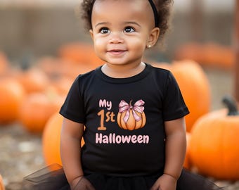 May include: A young child wearing a black t-shirt with the text "My 1st Halloween" and a pumpkin graphic with a pink bow. The child is also wearing a black tutu and a black headband. The background features several orange pumpkins.