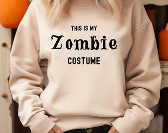 May include: A beige sweatshirt with the text "THIS IS MY Zombie COSTUME" in black. The sweatshirt has long sleeves and a crew neck. A pumpkin is visible in the background.