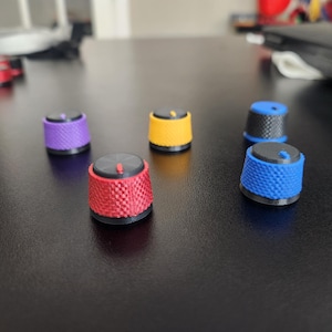May include: A collection of colorful, textured knobs in red, yellow, purple, blue, and black. Each knob has a black top with a small red indicator. The knobs are arranged on a dark surface.