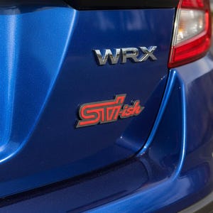 May include: A metallic blue car trunk featuring a chrome WRX emblem and a custom red and black badge that reads STI-ish. The surface has a glossy finish, and a portion of the red taillight assembly is visible on the upper right side of the frame.