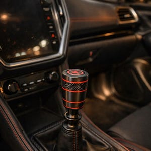 May include: Black and red car gear shift knob with a star emblem. The knob has a textured grip and red accent rings. The shift boot is black leather with red stitching, set in a black console.