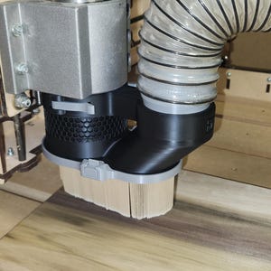 Puede incluir: Primer plano del husillo y el sistema de recogida de polvo de una máquina CNC. El husillo es plateado, con una cubierta negra de recogida de polvo y un accesorio de cepillo. Una manguera flexible está conectada a la cubierta. La máquina está trabajando sobre una superficie de madera.