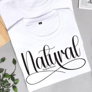 May include: Two white t-shirts with the word "Natural" in a cursive font. The text "Original Brand" is below the word "Natural". White trainers are visible in the background. The shirts are on a grey surface.