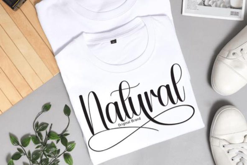May include: Two white t-shirts with the word "Natural" in a cursive font. The text "Original Brand" is below the word "Natural". White trainers are visible in the background. The shirts are on a grey surface.