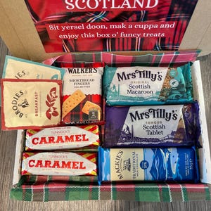 Pu&ograve; includere: Una scatola regalo in tartan rosso e verde piena di dolcetti scozzesi. La scatola contiene dita di shortbread, barrette di caramello, tavolette scozzesi e macarons scozzesi. La scatola ha il testo "A Taste of Scotland" in alto e "Sit yersel doon, mak a cuppa and enjoy this box o' funcy treats" in basso.