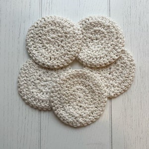 May include: Six cream-coloured, crocheted coasters are arranged on a white wooden surface. The coasters are circular, with a textured, handmade appearance. The neutral colour and simple design suggest a rustic or minimalist aesthetic.