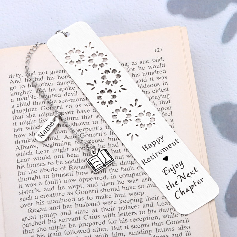 Puede incluir: Marcap&aacute;ginas de plata con un dise&ntilde;o recortado de fuegos artificiales. El marcap&aacute;ginas tiene un peque&ntilde;o amuleto de plata de un libro abierto unido a &eacute;l. El marcap&aacute;ginas tiene el texto "Happy Retirement" y "Enjoy the Next Chapter".