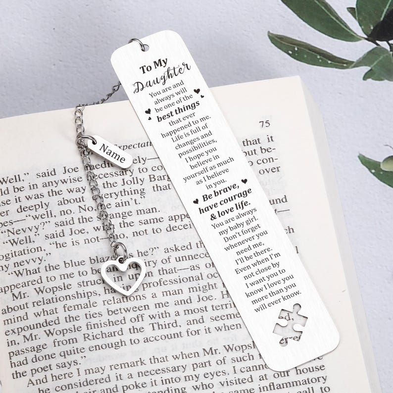 May include: A silver bookmark with a heart-shaped charm and a chain. The bookmark has a white background with black text that reads "To My Daughter" and includes a message about courage, love, and life. The bookmark is on a book with a white cover and black text.