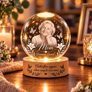 May include: A spherical glass ornament on a light wood base featuring an etched portrait of an older person, butterflies, and lilies. The text reads Mom, Forever Loved, and Butterflies appear when angels are near, illuminated by internal warm lights.